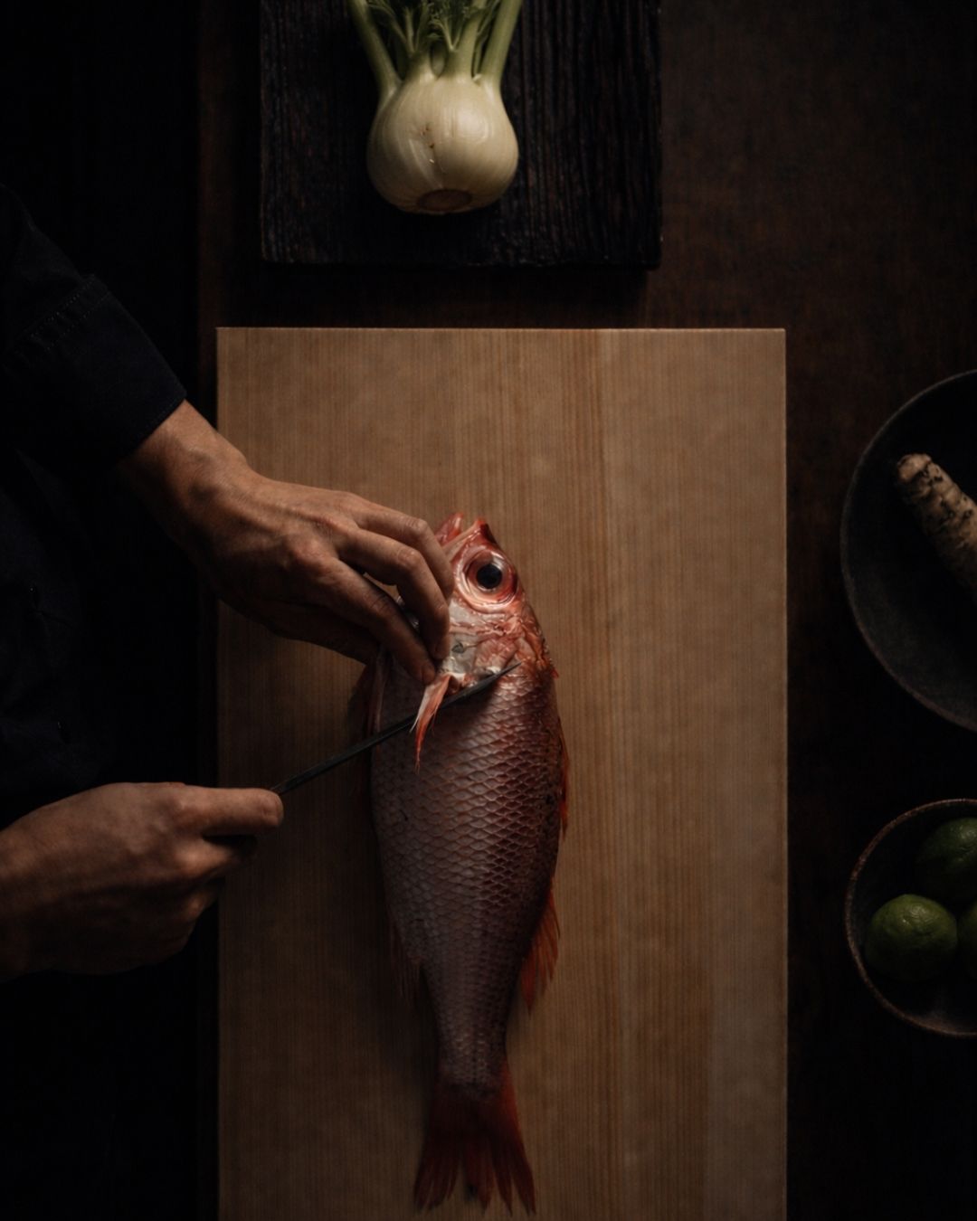 Person preparing a fish on a wooden cutting board with a dark background